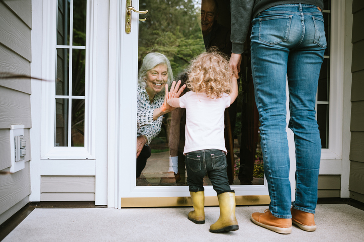 A mother stands with her daughter, visiting senior parents but observing social distancing with a glass door between them. The granddaughter puts her hand up to the glass, the grandfather and grandmother doing the same. A small connection in a time of separation during the Covid-19 pandemic.
